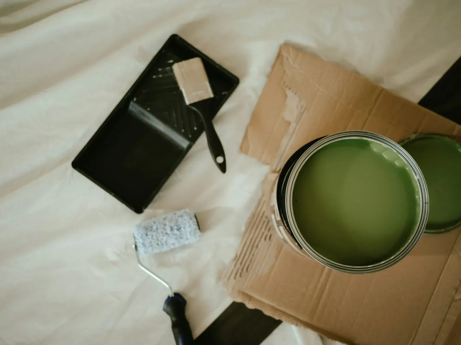An open tin of green paint on cardboard beside a tray, brush and roller sleeve, all set out on a drop sheet