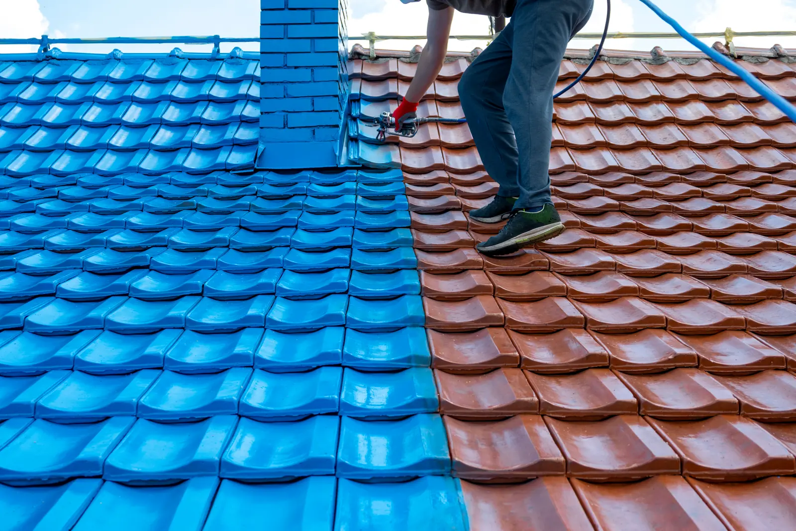 A roof half repainted - terracotta tiles on one side, fresh blue on the other, a painter spraying along the join