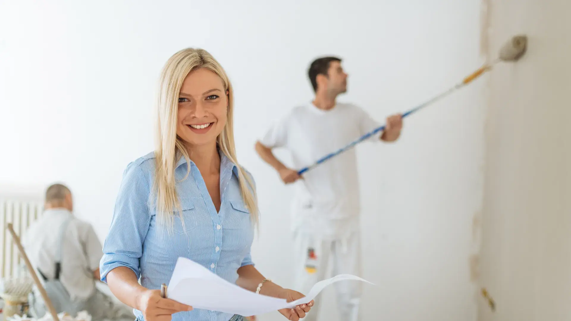 A homeowner with plans while a painter rolls the wall behind her